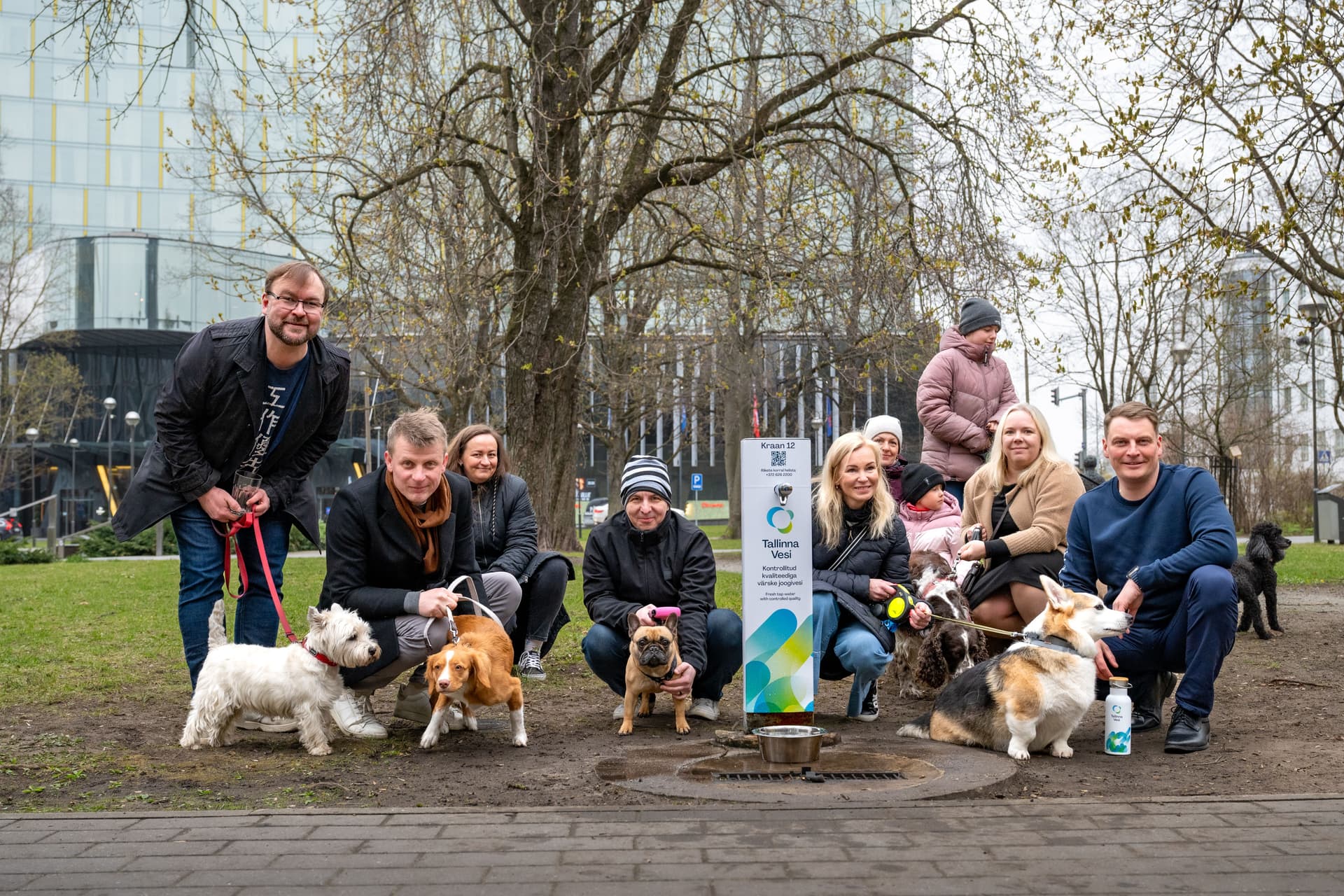 A diverse group of people and their dogs gather in a park around a pet-friendly sign.