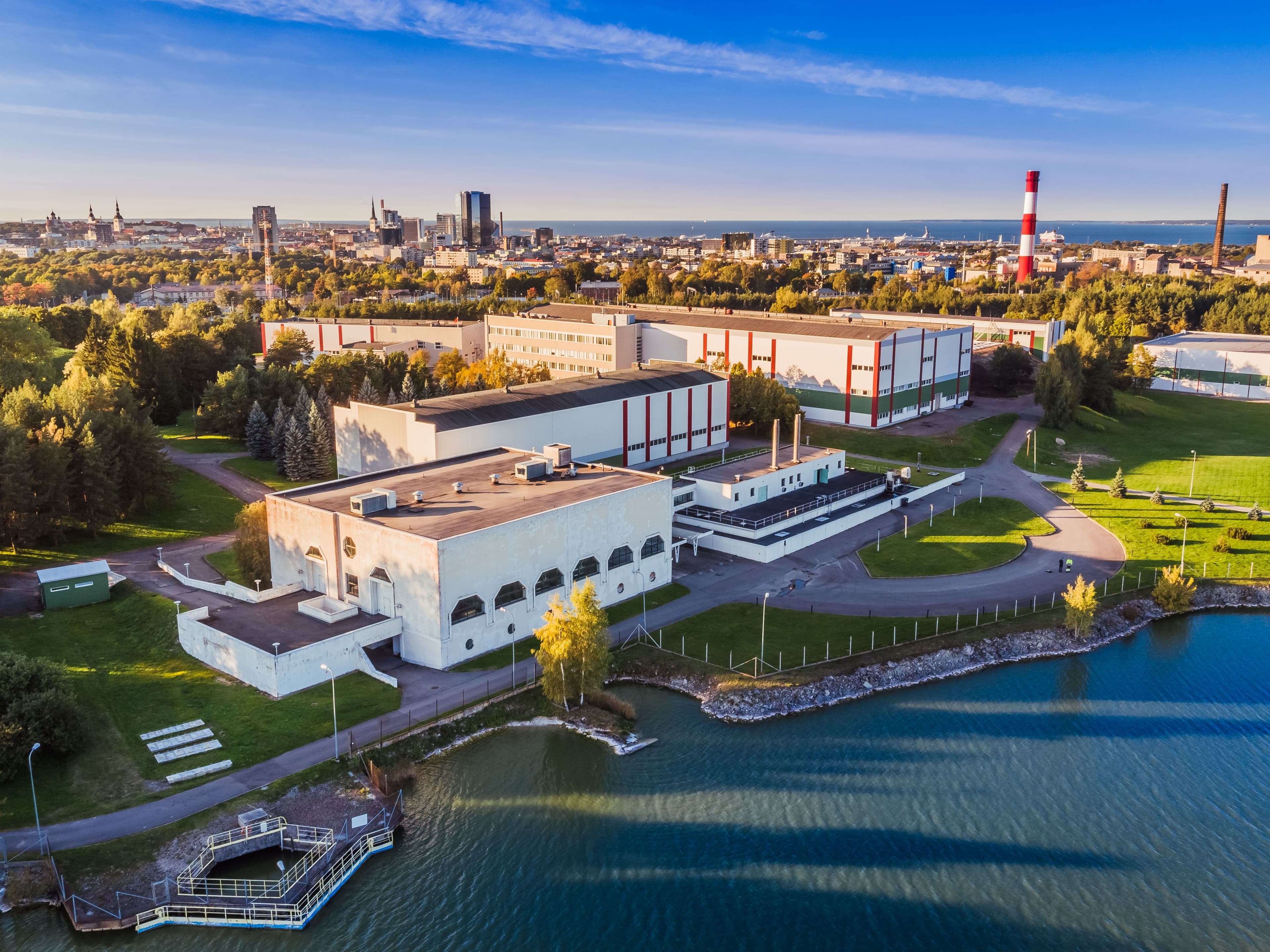 Aerial view of a large white building complex with a lake in the foreground and a city skyline in the background.