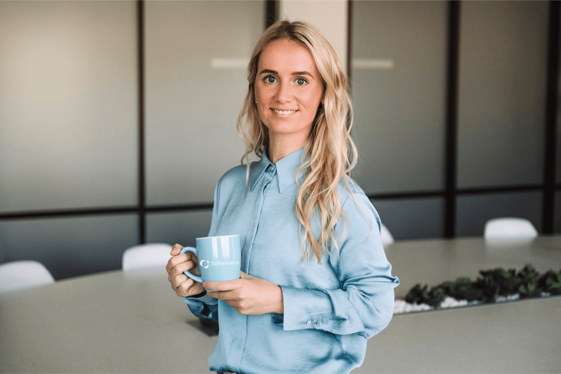 Young blonde woman in blue shirt holds a light blue mug in a modern office.