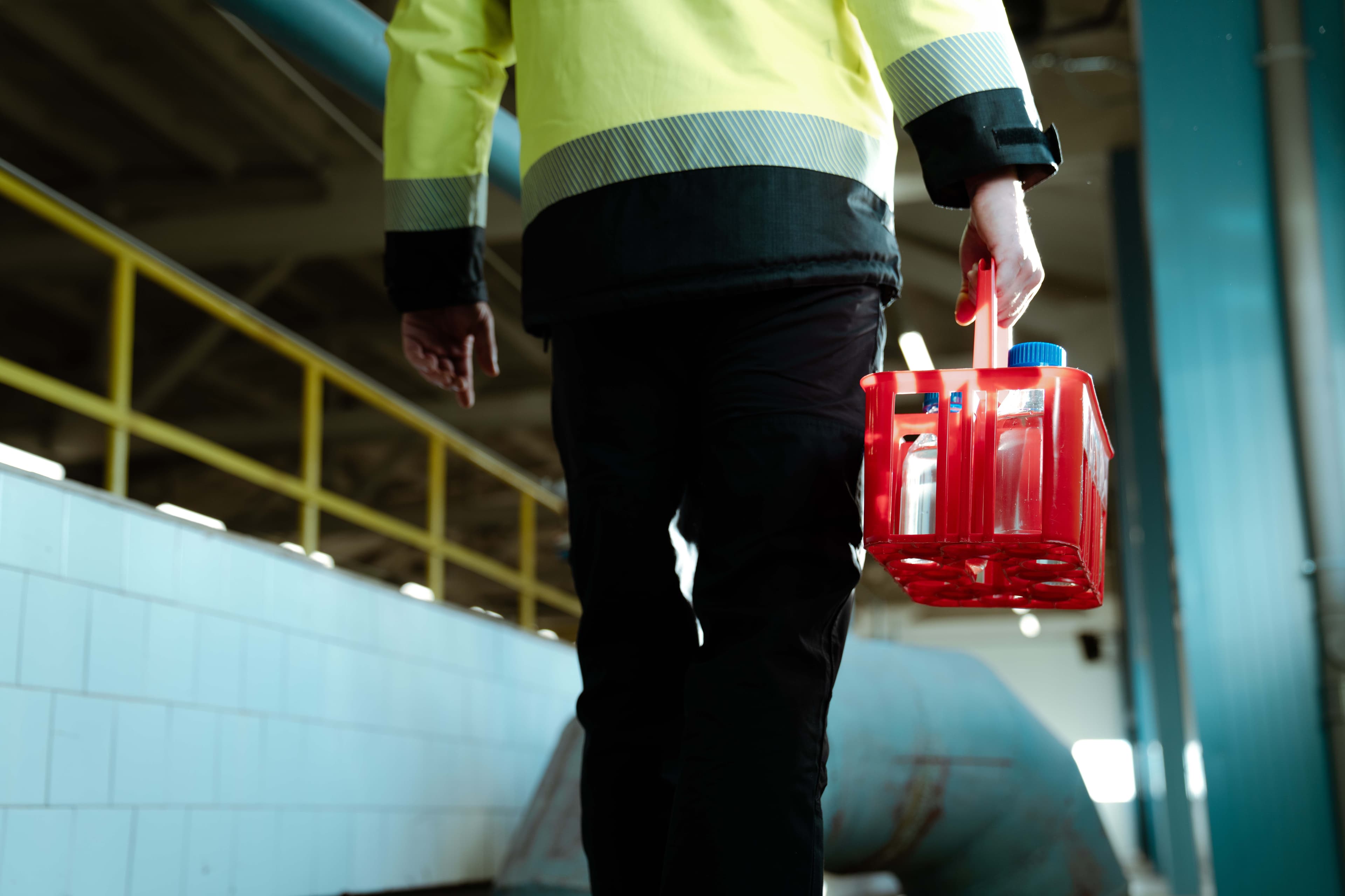 ööstushoones liikuv inimene, kellel on seljas kõrgnähtav kollane tööjope ja mustad püksid. Käes on punane plastikkast, milles on läbipaistvad pudelid siniste korkidega. Taustal on valge plaaditud sein, kollane käsipuu ja suur metalltoru.